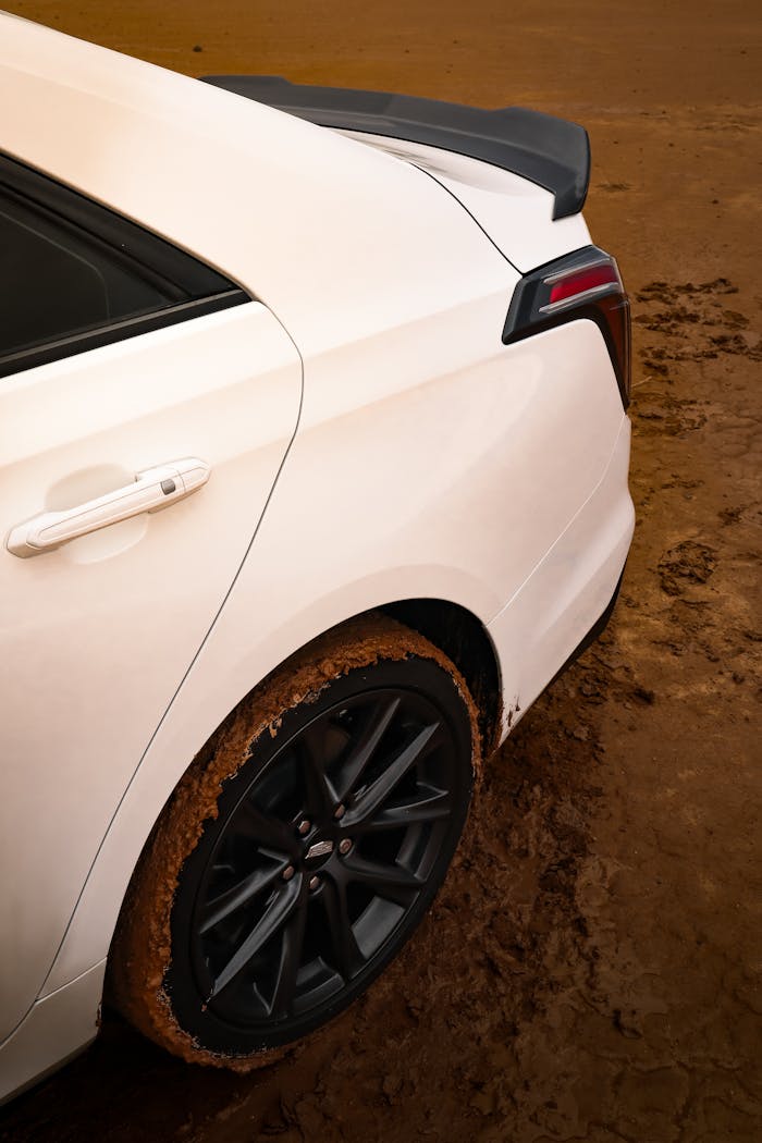 services-04 Close-up of a white car with muddy tires on a textured dirt road, showcasing rear design.